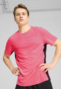 Camiseta atlética de manga corta rosa con paneles laterales negros, fabricada en tejido transpirable; cuenta con un pequeño logo plateado en el pecho.