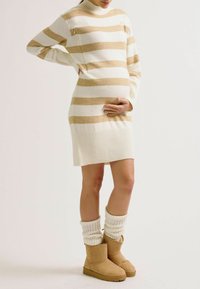 Robe-pull rayée en tricot, crème et marron clair, avec col montant, ourlet côtelé et fermetures éclair à l'avant. Associée à des bottes beige et des poignets crème.