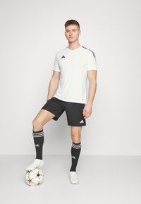 Camiseta atlética blanca con rayas negras, combinada con pantalones cortos negros, calcetas negras hasta la rodilla con rayas blancas y zapatillas de fútbol blancas.