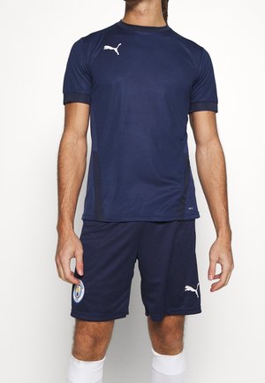Mand iført navyblå Puma sportstrøje og shorts med Manchester City-logo på venstre ben af shortsene og hvide sokker.