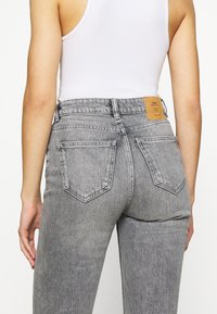 Persona con jeans de mezclilla gris de cintura alta y bolsillos traseros, con una etiqueta de marca color beige, combinados con una blusa sin mangas blanca.