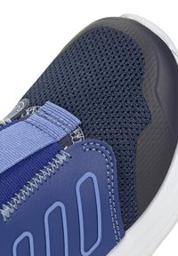 Primer plano de la puntera de una zapatilla azul con parte superior de malla, superposiciones sintéticas, cinturón elástico y suela de goma blanca.