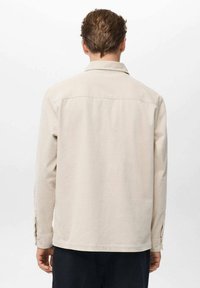 Camisa de pana beige con mangas largas, puños abotonados y corte recto. Presenta un yugo trasero y una textura sutil.