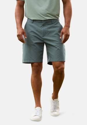 Hombre con pantalones cortos verde azulado, camisa verde claro y zapatillas blancas, de pie con las manos relajadas a los lados contra un fondo liso.