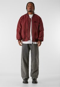 Bordó bomber bunda s žebrovanými manžetami, zipem a postranními kapsami. V kombinaci s širokými šedými kalhotami a černými botami. Pod ní je obyčejné bílé tričko.