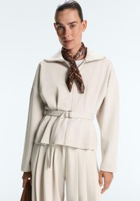 Ženska, oblečena v kremno suknjo z zaponko in plisirane hlače, z rjavo vzorčasto rutko okoli vratu, drži rjavo teksturirano torbico.