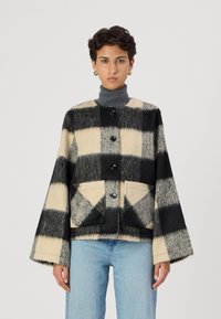 Veste en polaire avec un motif à carreaux noir et crème, dotée d'un col rond, de longues manches évasées, de cinq boutons et de deux poches avant.