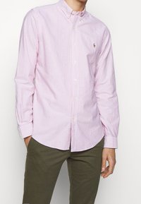 Rosa och vitrandig skjorta med knappar, spridd krage och långa ärmar, med en liten broderad logotyp på vänster bröst.