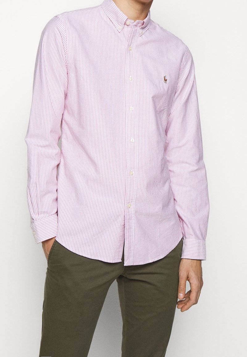 Rosa och vitrandig skjorta med knappar, spridd krage och långa ärmar, med en liten broderad logotyp på vänster bröst.