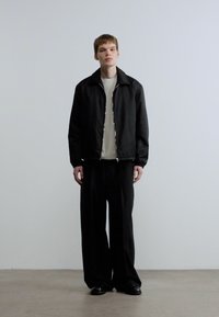 Νεαρός άνδρας που στέκεται μπροστά σε λευκό τοίχο, φοράει μαύρο μπουφάν με φερμουάρ, λευκό πουκάμισο, φαρδιά μαύρα παντελόνια και μαύρα παπούτσια με ουδέτερη έκφραση.