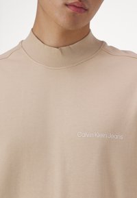 Felpa beige a collo alto realizzata in tessuto liscio, con un sottile motivo testurizzato e ricamo bianco "Calvin Klein Jeans" sulla parte anteriore.