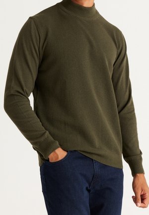 Mann trägt einen olivgrünen, strukturierten Pullover mit langen Ärmeln, gerippten Bündchen und Kragen, kombiniert mit dunkelblauen Jeans, eine Hand in der Tasche.