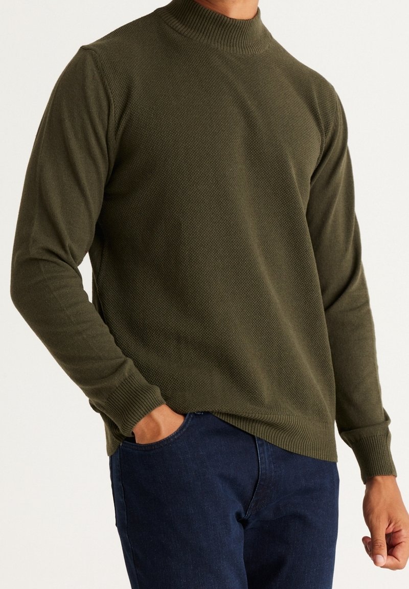Homme portant un pull texturé vert olive à manches longues avec poignets et col côtelés, associé à un jean bleu foncé, main dans la poche.
