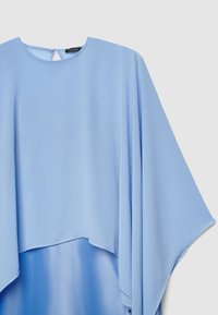 Light blue flowing blouse with wide sleeves and a round neckline, featuring a small back button closure and layered hem.