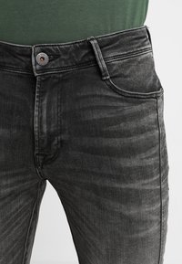 Jeans de mezclilla negra con un lavado desgastado, que cuentan con un bolsillo frontal, cierre de botón metálico y sutiles detalles de costura. Textura de tela ligera.