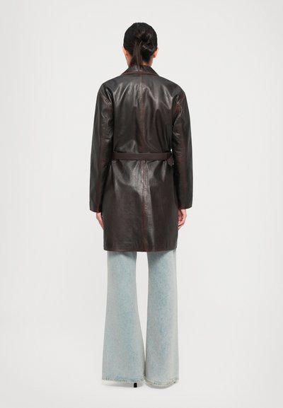 Donna con i capelli scuri raccolti in una coda di cavallo, indossa un cappotto in pelle marrone scuro con cintura e jeans larghi di colore blu chiaro, in piedi davanti a uno sfondo semplice.