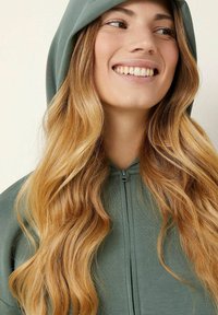 Felpa verde con cappuccio e zip frontale, caratterizzata da una texture morbida e una vestibilità rilassata, indossata da un individuo con lunghi capelli mossi.