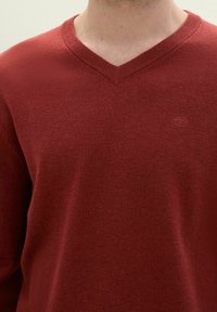 Raudonas V-kaklo megztinis, pagamintas iš minkštos medžiagos su lygiu paviršiumi. Turi ribotu kaklo apvadu ir mažą, iškilnią logotipo etikete krūtinėje.