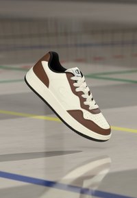 Λευκό και καφέ δερμάτινο αθλητικό παπούτσι με διάτρητο μπροστινό μέρος, μαύρες λεπτομέρειες και αντίθετο λογότυπο· καουτσούκ εξωτερική σόλα με υφή για καλύτερη πρόσφυση.