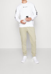 Vit sweatshirt med svart logotyp på bröstet och ärmarna, beige sweatpants och vita sneakers. Håller en genomskinlig vattenflaska.