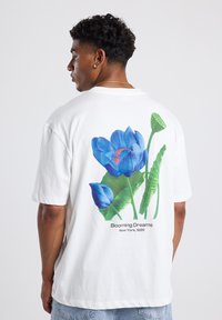 Camisa de algodón blanca con un vibrante gráfico floral azul en la parte posterior, con hojas verdes y el texto "Sueños en Flor, Nueva York, 1989."