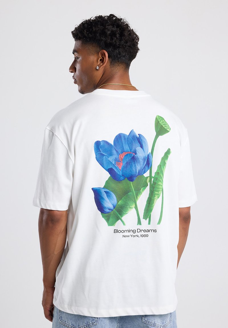 Camisa de algodón blanca con un vibrante gráfico floral azul en la parte posterior, con hojas verdes y el texto "Sueños en Flor, Nueva York, 1989."