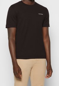 Černé bavlněné tričko s krátkým rukávem, kulatým výstřihem a malým bílým logem Calvin Klein na hrudi. Jednoduchý, jednobarevný design.