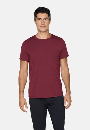 Mand, der står med fronten mod kameraet iført en kortærmet vinrød T-shirt med brystlomme og sorte bukser, neutralt baggrund.