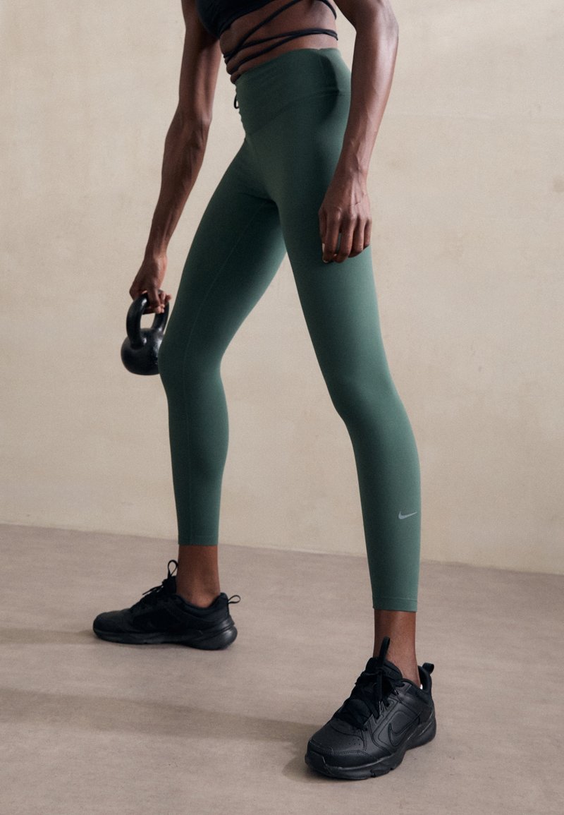 Leggings verdes de cintura alta con una textura suave y un ajuste que realza la figura, combinados con zapatillas deportivas negras. Sosteniendo una kettlebell negra.