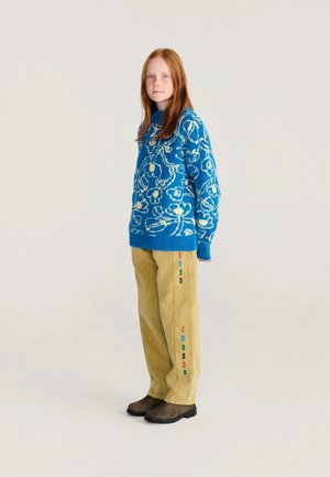 Enfant aux longs cheveux roux portant un pull bleu à motifs floraux, un pantalon jaune avec un texte coloré sur le côté et des bottes marron, debout devant un fond uni.