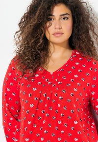 Rotes Oberteil mit langen Ärmeln, V-Ausschnitt und verspieltem Blumenmuster in Schwarz und Weiß. Der Stoff wirkt weich und leicht.