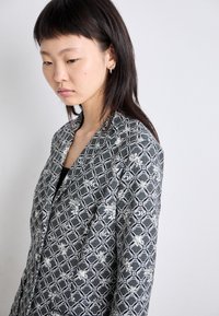 Schwarz-weiß gemusterte Jacke mit hexagonalen Formen und floralen Akzenten. Glattes Material, offenes Frontdesign und kragenloser Stil.