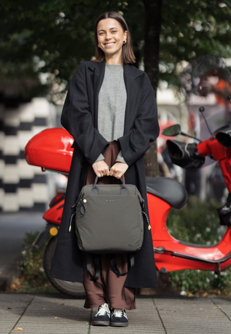 Jeune femme en long manteau sombre et baskets souriante, tenant un sac gris devant un scooter rouge sur un trottoir en ville.