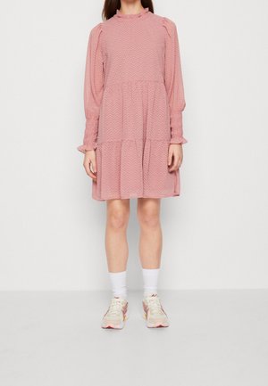 Frau steht und trägt ein rosa Langarmkleid mit Rüschen, weiße Socken und weiße Sneakers vor einem weißen Hintergrund.