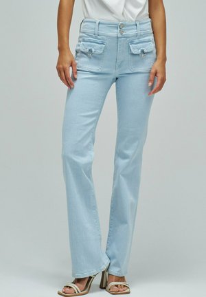 Mujer con jeans de tiro alto azul claro con bolsillos delanteros con solapa y cierre de doble botón, combinados con sandalias beige de tacón.