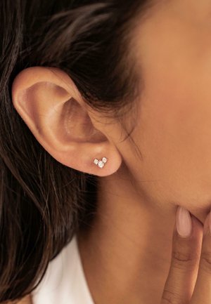 Diamantsteckerohrringe mit einem Design aus drei Steinen, mit einer Metallfassung, auf einer Nahaufnahme des Ohrs platziert, die die Kontur des Ohrs hervorhebt.