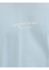 Light blue cotton t-shirt featuring the text "Originals Studio" in white, with additional text underneath. Smooth texture and casual design.