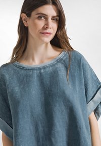 Top in lino azzurro chiaro con scollo ampio e maniche corte cadenti. Il tessuto ha una finitura leggermente strutturata e una silhouette casual.