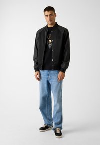 Zwarte jas met een gladde textuur en drukknoopsluiting, gecombineerd met een zwart T-shirt met print en lichtblauwe jeans met rechte broekspijpen, gedragen met zwarte sneakers.