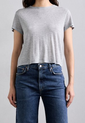 Kvinde iført en ensfarvet lysegrå kortærmet crop-top og højtaljede blå cowboybukser, der står med afslappede arme.