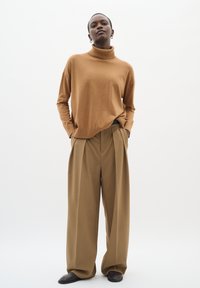 Brun turtleneck-trøje med løs pasform, kombineret med beige bukser med høj talje og vide ben. Modellen har mørke sko på og står mod en ensfarvet baggrund.