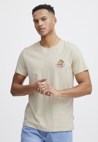 Beige bomulds t-shirt med korte ærmer, rund halsudskæring, med en orange grafik og grønne bladmønstre broderet samt tekst nedenfor.