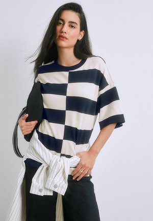 Fiatal nő hosszú, sötét hajjal, fekete-fehér kockás inget visel, fehér csíkos dzsekit kötött meg, és fekete táskát hord.