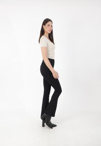 Top ajustado beige, pantalones acampanados negros y botas de tacón cuadrado negras. La modelo está de lado frente a un fondo blanco.