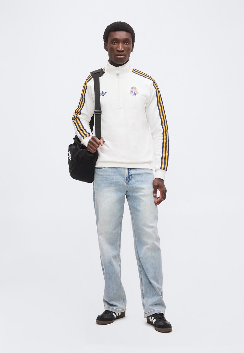 Homme portant une veste de sport blanche avec des rayures jaunes et bleues, un jean bleu clair, des baskets noires, et tenant un sac à bandoulière noir Adidas.