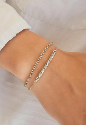 Due braccialetti delicati a catena d'argento al polso sopra una camicia chiara a maniche lunghe. Uno dei braccialetti ha un design intrecciato e testurizzato.