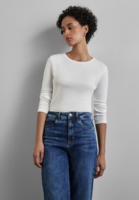 Weißes Langarmhemd mit rundem Halsausschnitt, kombiniert mit hochgeschnittenen dunkelblauen Jeans. Glatte Stoffoberfläche, klassischer Schnitt, keine sichtbaren Muster.