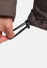 Giacca isolante marrone con una mano che regola un cordino all'orlo, dotata di corda elastica nera e manettino per Stringere.
