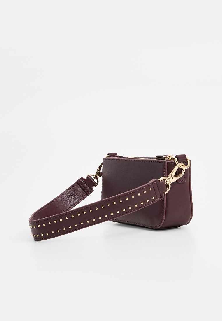 Kleine bordeauxrote Lederhandtasche mit goldenen Beschlägen, einem nietenbesetzten Tragegurt, Reißverschlussverschluss und rechteckiger Form. Glatte Textur mit minimaler Detailverarbeitung.