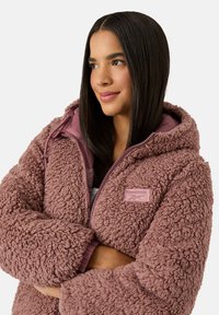 Kurtka polarowa w kolorze pudrowego różu z fakturowaną warstwą zewnętrzną, zamkiem błyskawicznym i kapturem. Na piersi znajduje się naszywka z logo Reebok.